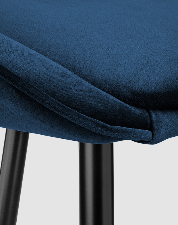 Close-up corner view of a barstool seat. Its blue velvet upholstery has a neat piping trim; black steel legs are underneath. Close-up corner view of a barstool seat. Its blue velvet upholstery has a neat piping trim; black steel legs are underneath.