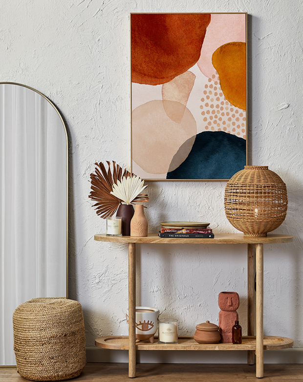A round timber console table is styled against a textured white wall and below colourful abstract artwork.