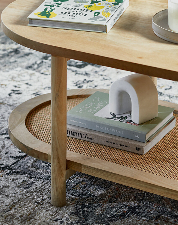 A zoomed-in view of an oval-shaped coffee table, with books on both the mango wood top and the rattan lower shelf.