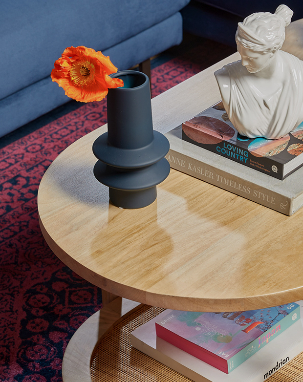 Angled bird's-eye view of a timber coffee table. An orange flower in a vase, books, and a white bust are styled on top.