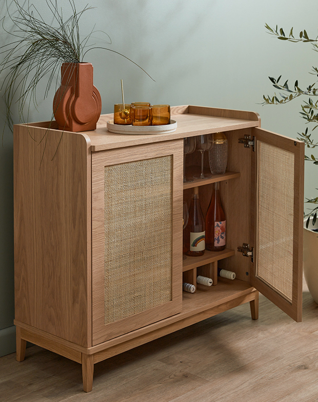 Against a mint green wall, a buffet cabinet has one of its doors open, revealing bottles of wine and barware stored inside.