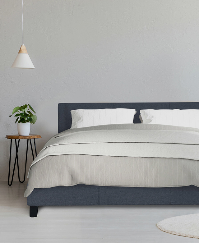 The bed is made with grey striped sheets. A peg leg side table is next to it with a plant on top, & a pendant hangs over it. The bed is made with grey striped sheets. A peg leg side table is next to it with a plant on top, & a pendant hangs over it.