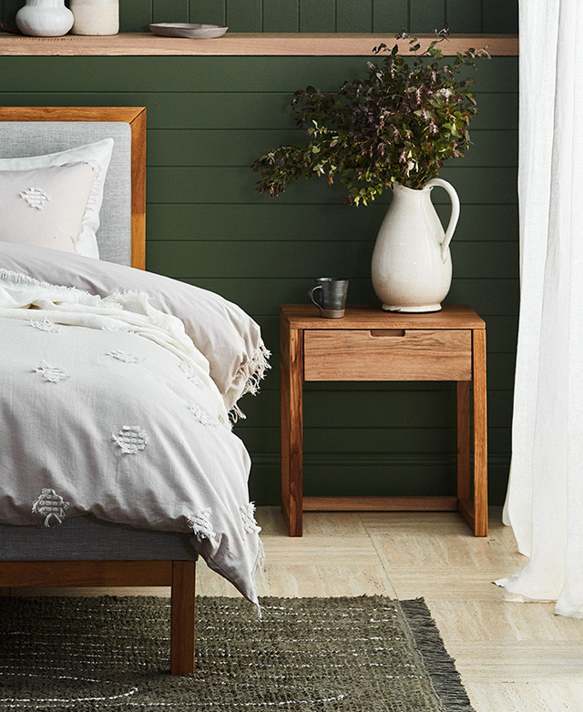 The table is in a bedroom, next to a bed. The panelled wall it's in front of is green, and a jug of green foliage is on top.