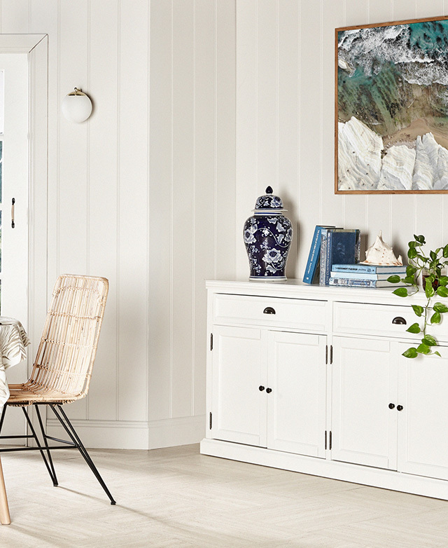 The sideboard is styled in a light-filled, white room with panelled walls, light flooring and a rattan chair.