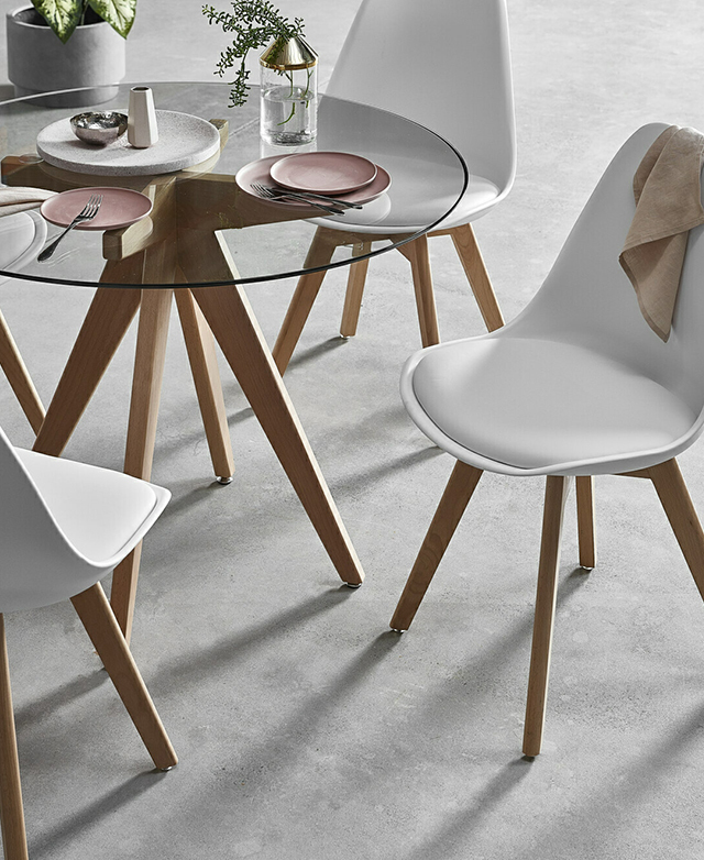 Three chairs are positioned around a glass-top round table. The floor is cool concrete. On the table is pink crockery.