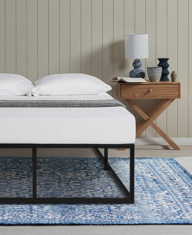 A side table with decor & a lamp is next to the bed. A patterned rug is beneath. The generous space under the bed is shown.