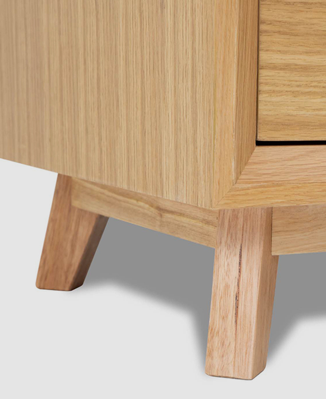 Angled view of the bedside table base. It features a thick plinth and solid timber legs that have been splayed for stability.