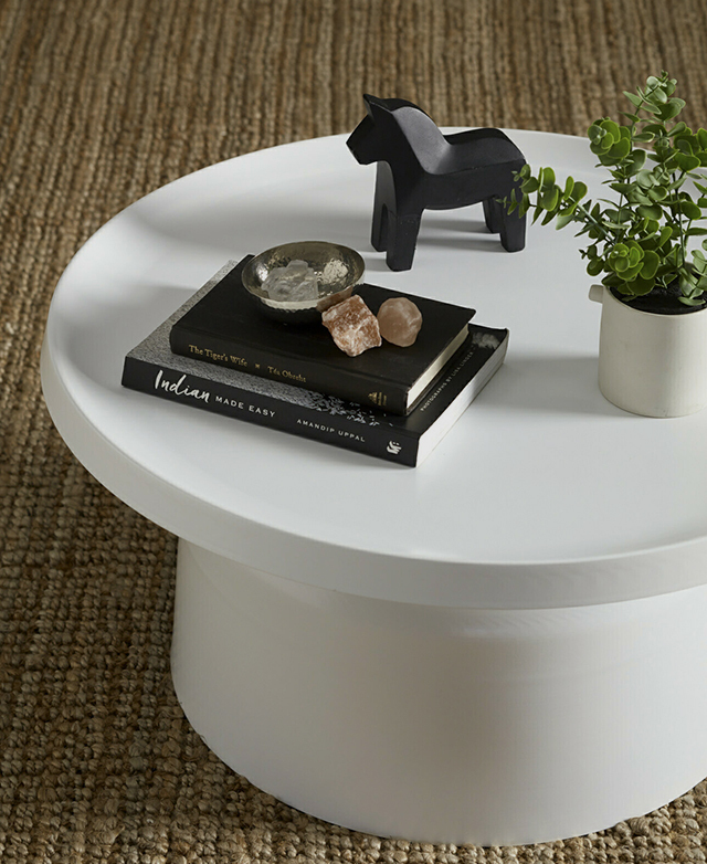 A cropped image shows a white coffee table complete with books, crystals, a plant and decor, all on top of a jute rug. A cropped image shows a white coffee table complete with books, crystals, a plant and decor, all on top of a jute rug.