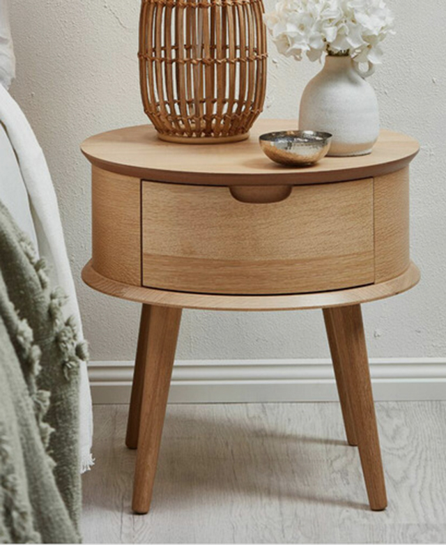 A rattan cage lamp, white ceramic vase with white flowers, and metal pinch bowl is on top of the side table.