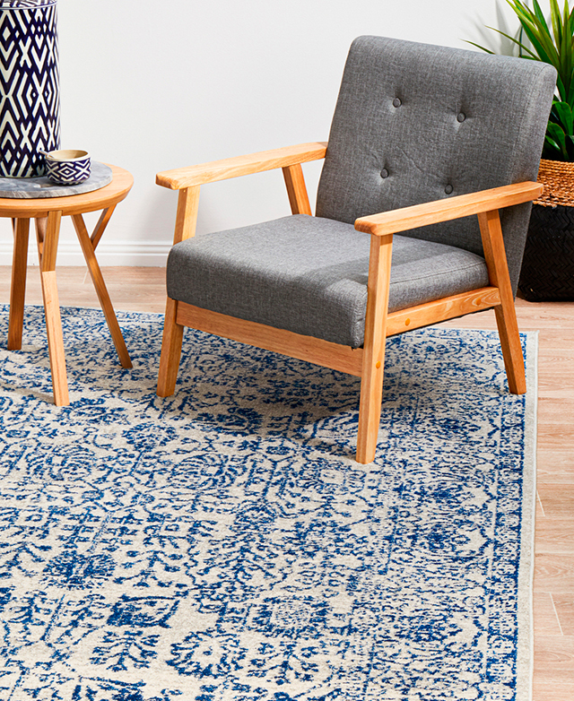 A polypropylene rug with a blue floral pattern on a white background. The rug is styled under a timber chair and side table.