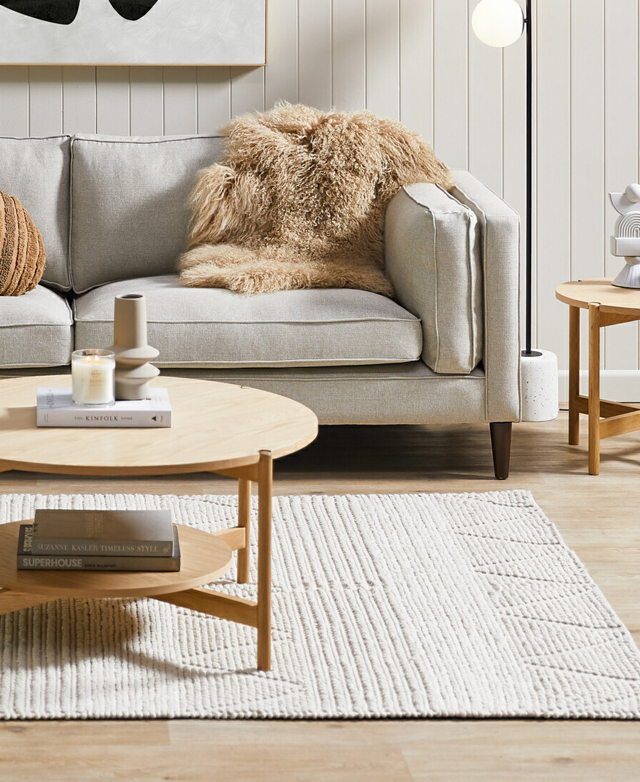 A round, blonde wood coffee table with two levels sits on top of the rug, which is positioned near a neutral-toned sofa.