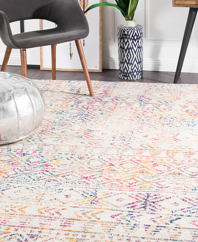 A rainbow-coloured rug with a geometric diamond pattern on top of dark timber flooring and underneath sparse furniture.