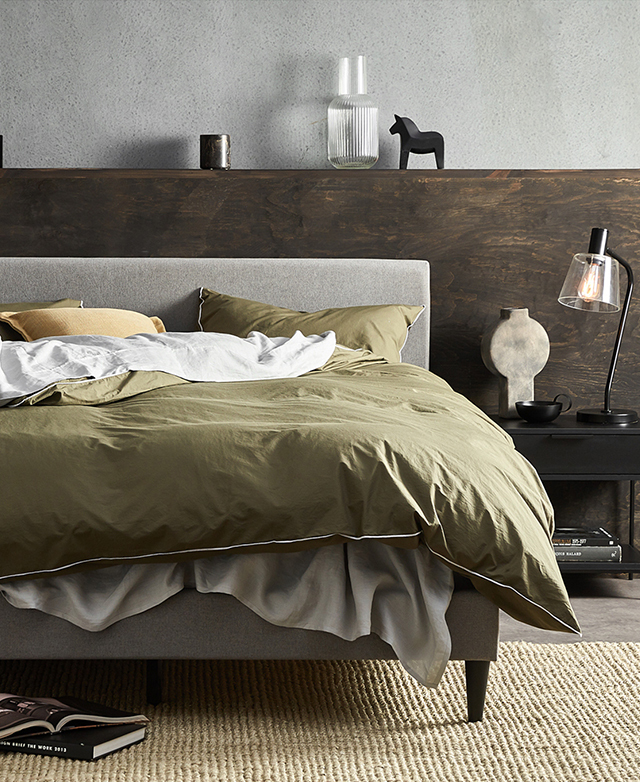 The bed is in front of a dark brown half-wall and on a jute rug, dressed in complementary green and grey sheets. The bed is in front of a dark brown half-wall and on a jute rug, dressed in complementary green and grey sheets.