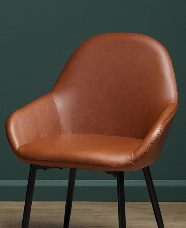 A chair with brown faux leather upholstery. Lightly padded, the seat features mid-height armrests that are angled outwards. A chair with brown faux leather upholstery. Lightly padded, the seat features mid-height armrests that are angled outwards.
