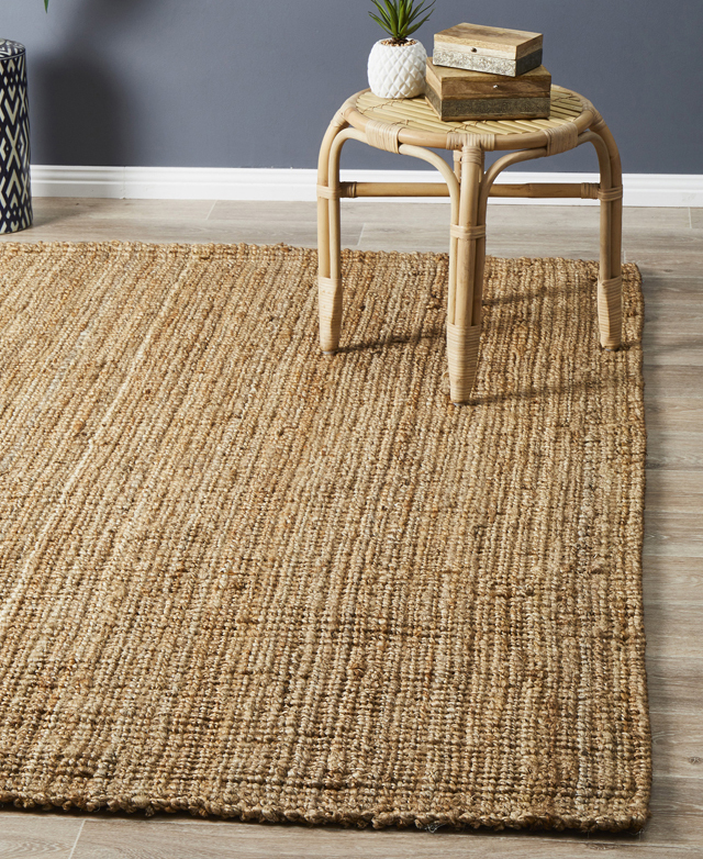 The jute rug is in a room with a blue-grey wall & white skirting board behind, & a round rattan stool with adornments on top.