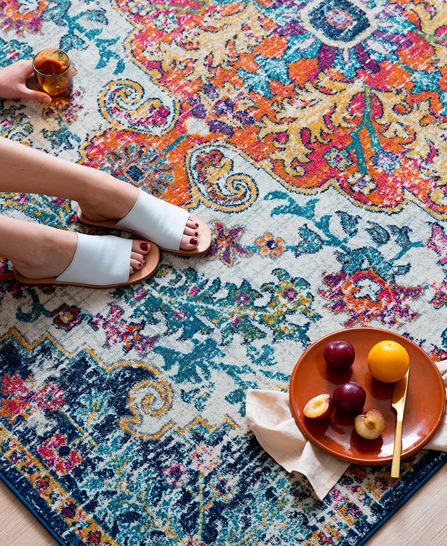 A person with white slides sits on the rug, next to a bowl of fruit and an amber glass.