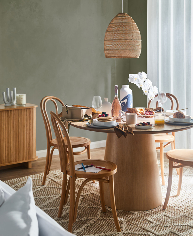 Four natural chairs are around a pedestal table. Various breakfast items are on the table, such as waffles and orange juice.