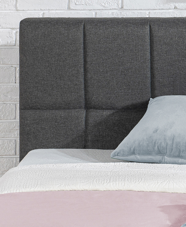 A close-up of the grey polyester bedhead shows the quilted stitch details and lofty padding. A close-up of the grey polyester bedhead shows the quilted stitch details and lofty padding.