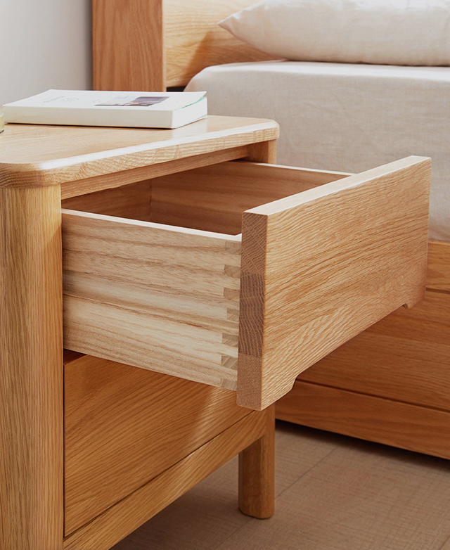 A three-quarter angle of the wood bedside table shows the top drawer open. It's in front of a bed, & has a white book on top.