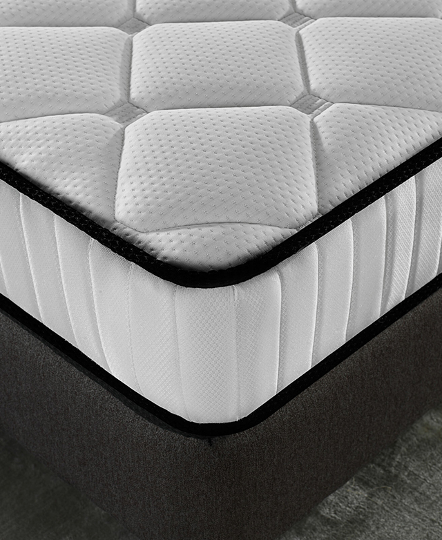 A corner of the mattress is displayed close up. Its black piped trims frame the white top and middle section.
