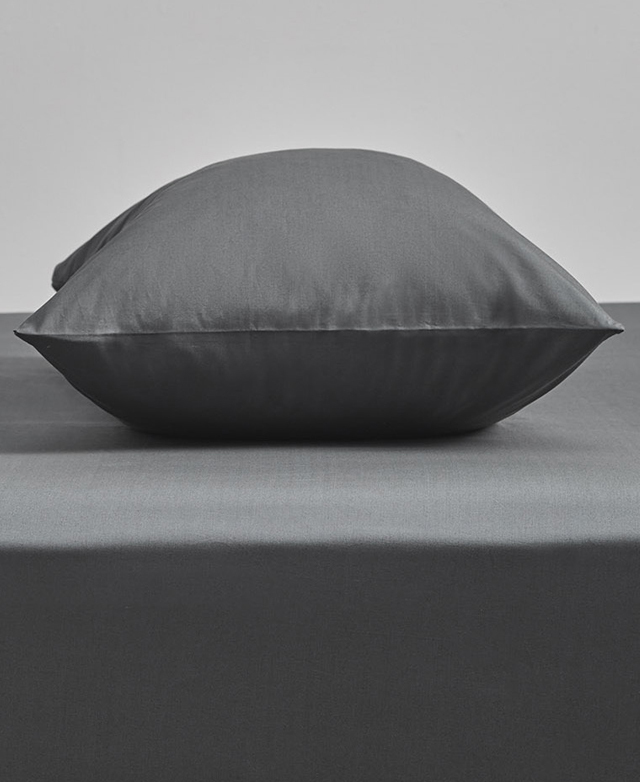 Eye-level side view of a plump pillow encased in a 100% cotton pillowcase on top of a bed with a grey fitted sheet.