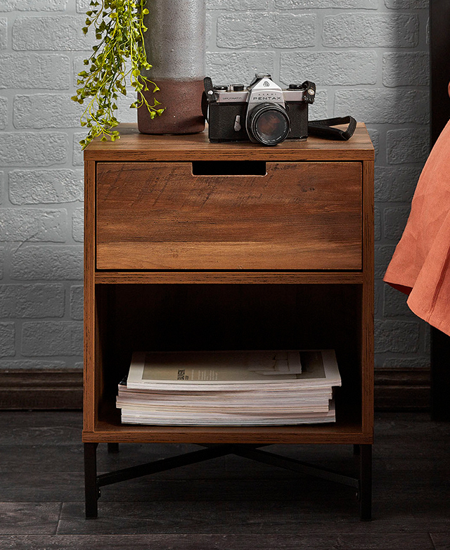 The dark wood bedside table has a stack of magazines on the lower shelf, and a tall vase with leafy greenery on the table top.