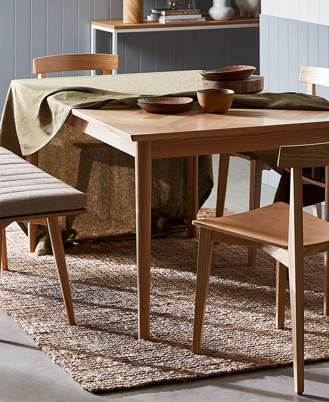 The table is styled on a jute rug, with seating positioned around it. An olive table cloth is partially draped over the top. The table is styled on a jute rug, with seating positioned around it. An olive table cloth is partially draped over the top.