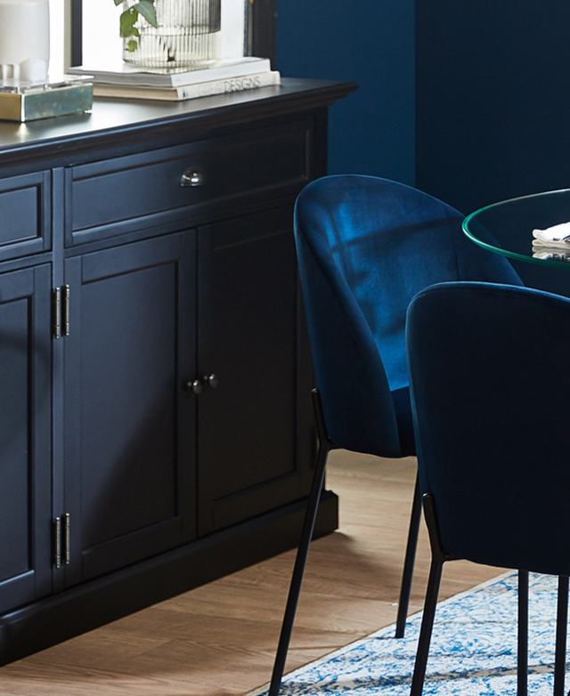 The sideboard is in a dining room setting, with jewel-like blue velvet chairs and a glass dining table placed nearby.