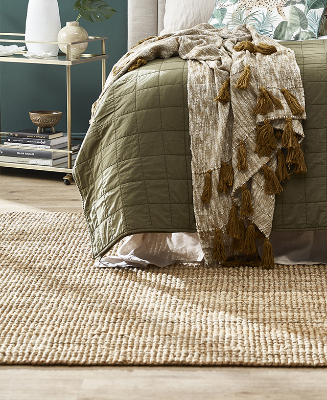 The jute rug is positioned beneath a bed, which is dressed in layers of earthy green tones.