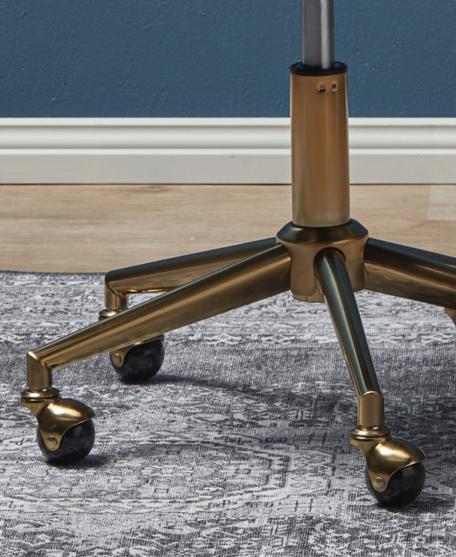 The brass five-point star base of a chair on top of a grey patterned rug. Round castor wheels are at the end of each point.