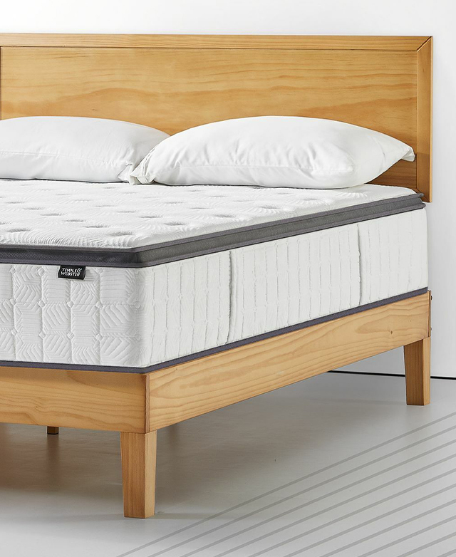 A three-quarter angle displays the undressed mattress on a simple yellow-toned wooden bed. Two white pillows are on top.
