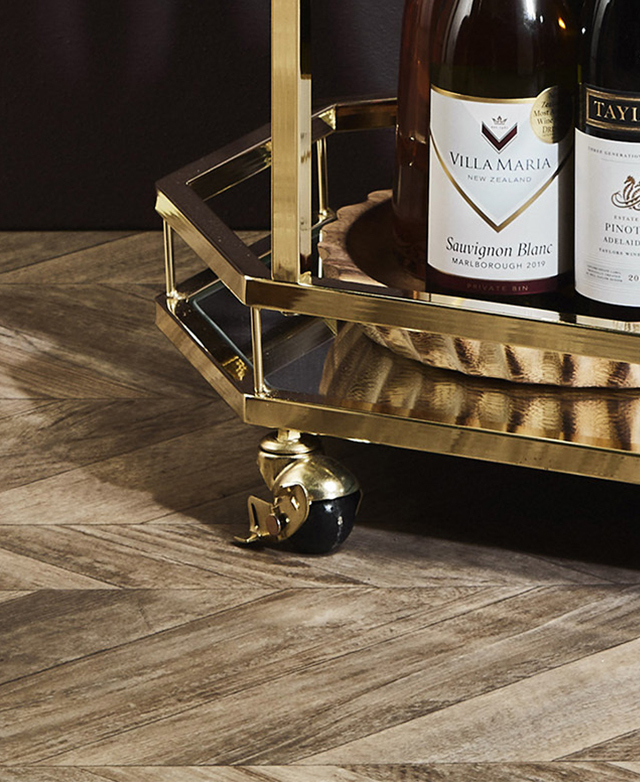 Up close is a golden, lockable castor wheel at the base of the bar cart. The polished bottom shelf holds wine bottles.