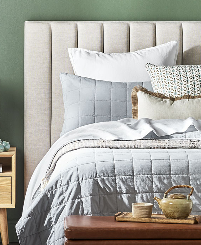 A bed is shown fully dressed, with layers of cool-toned bedding and cushions positioned in front of the bedhead.