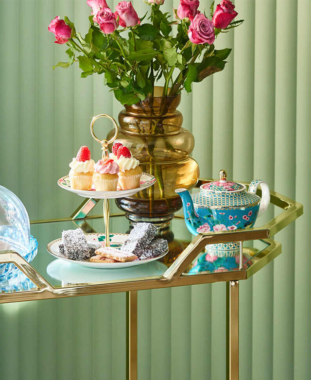 Near a green fluted wall, a high tea is set up on the cart. It holds a tiered cake stand with treats, a teapot & pink roses.