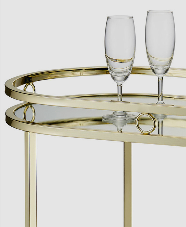The top of an oval, gold-framed bar cart is shown, with two champagne flutes resting atop the mirrored glass shelf. The top of an oval, gold-framed bar cart is shown, with two champagne flutes resting atop the mirrored glass shelf.