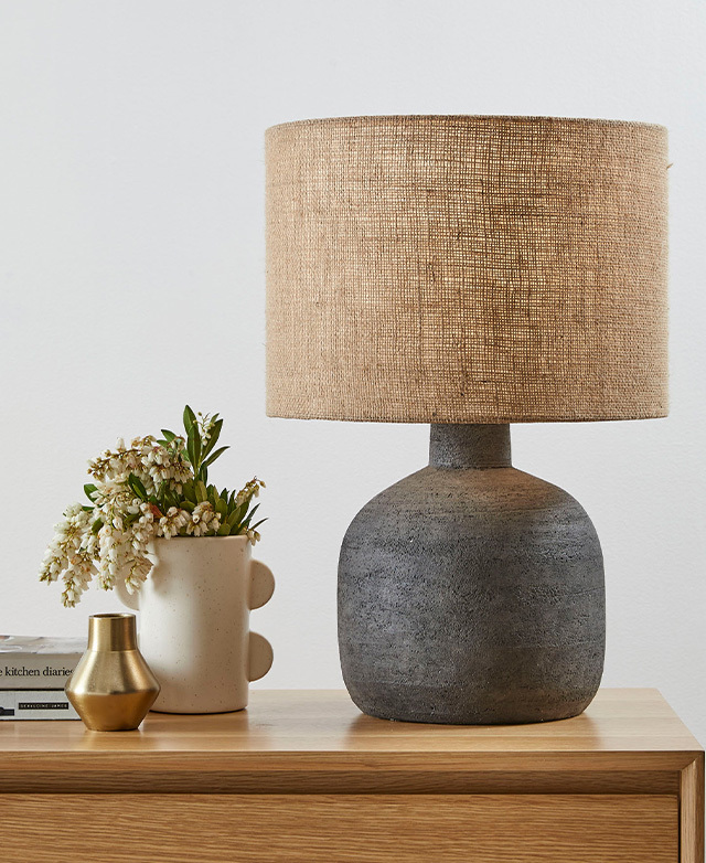A lamp with a textured hessian lampshade and black terracotta base is styled on a timber dresser next to a vase of flowers.