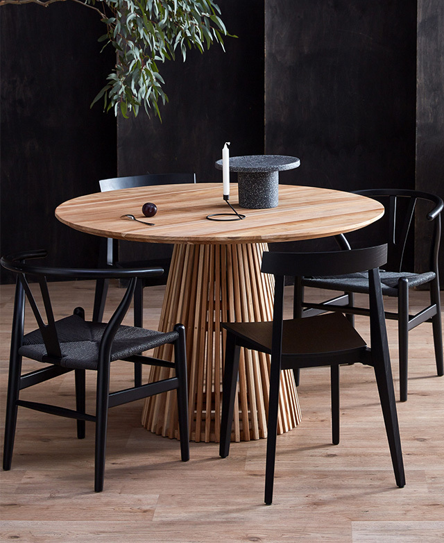 A teak wood dining table styled with black Wishbone dining chairs in a bold dining room. A leafy plant is in the background.