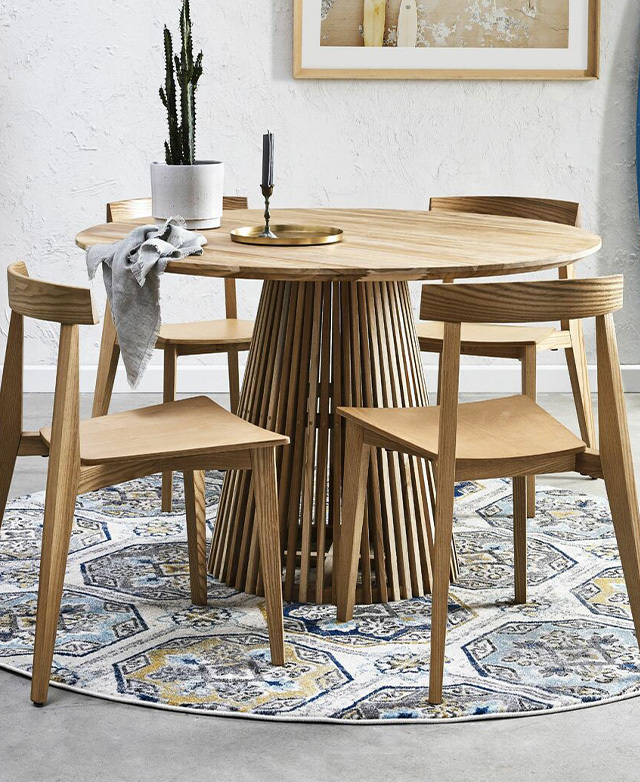 A round dining table and minimalist timber dining chairs styled above a circular rug with a geometric pattern.