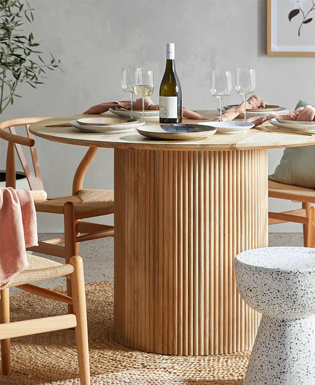 The dining table is dressed with crockery, pink napkins, and glasses, and styled with wishbone chairs and a stool.