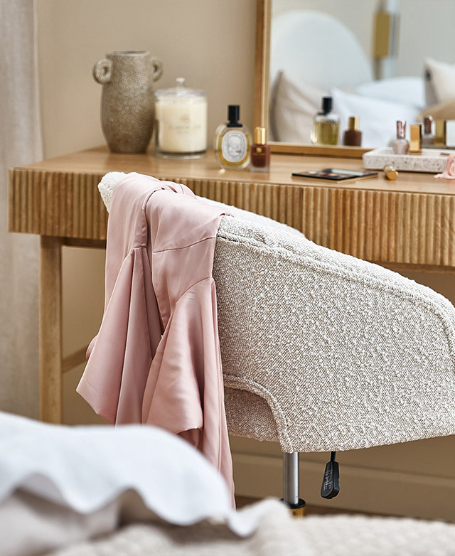 Perfume bottles and make-up scattered on top of a mango wood table. A boucle armchair is depicted in the foreground.
