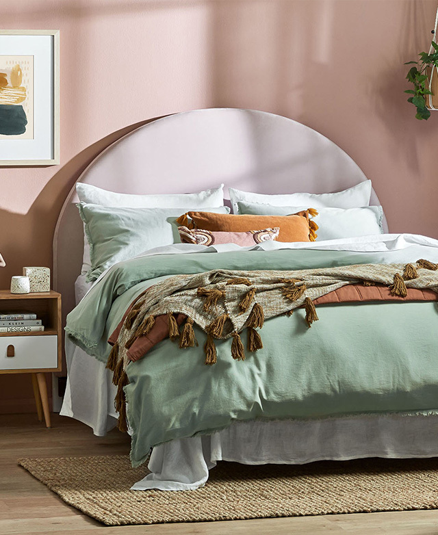 An arch bedhead with blush pink velvet upholstery is positioned behind a bed dressed in white and sage green bedding.