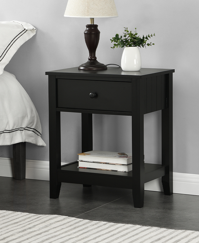 A black side table is positioned next to a bed, with a lamp and plant on top, and stack of books on the shelf below.