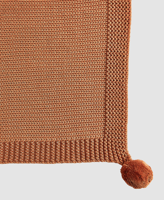 Close-up of a rust-coloured 100% cotton throw highlights its cosy knitted texture and soft pom pom accent on the corner.