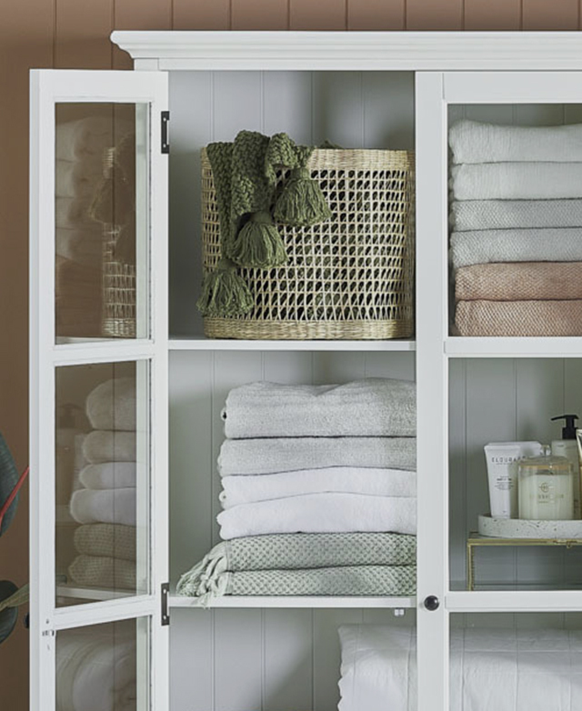 A door is fully open to show a woven rattan basket, which holds a tasselled green towel. Other bathware is on the shelves.