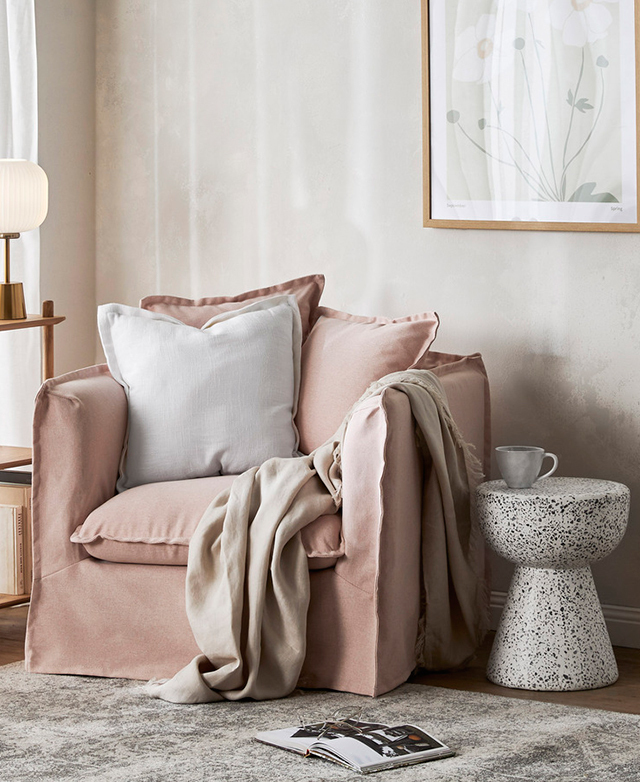 Light softly fills the room that contains the armchair. It has an additional cushion, and a linen blanket draped over the arm.