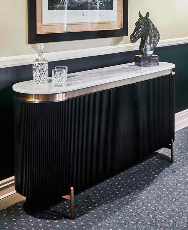 An oval buffet table with a fluted black body. A decorative horse statue and whiskey decanter are on the real marble top.