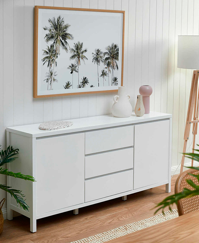 A white rectangular sideboard buffet is styled against a white panelled wall. Landscape artwork of palm trees hangs above.