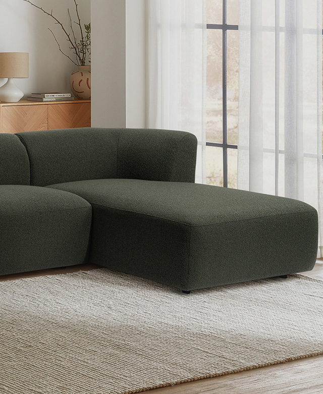 The simple green chaise end of the sofa is on a cream rug, running alongside a large window with sheer curtains. The simple green chaise end of the sofa is on a cream rug, running alongside a large window with sheer curtains.