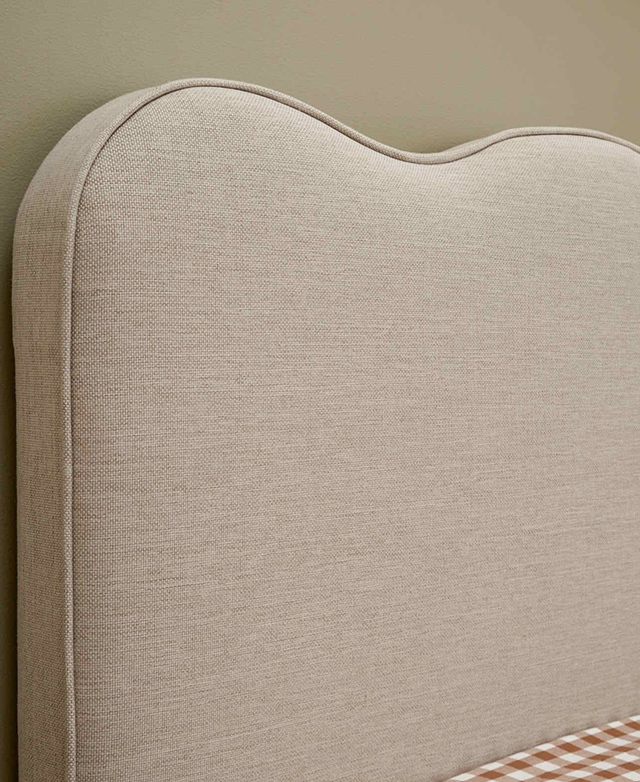Closely cropped three-quarter angle of the bedhead, which has a wavy top shape upholstered in taupe polyester. Closely cropped three-quarter angle of the bedhead, which has a wavy top shape upholstered in taupe polyester.