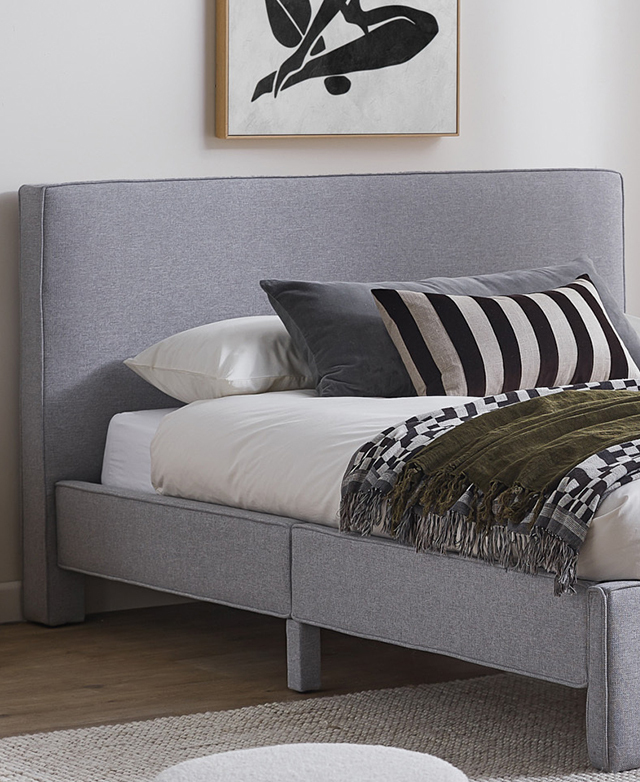 The fully made grey bed features cream, grey, and green bedding in a white room with a monochrome artwork hanging above.